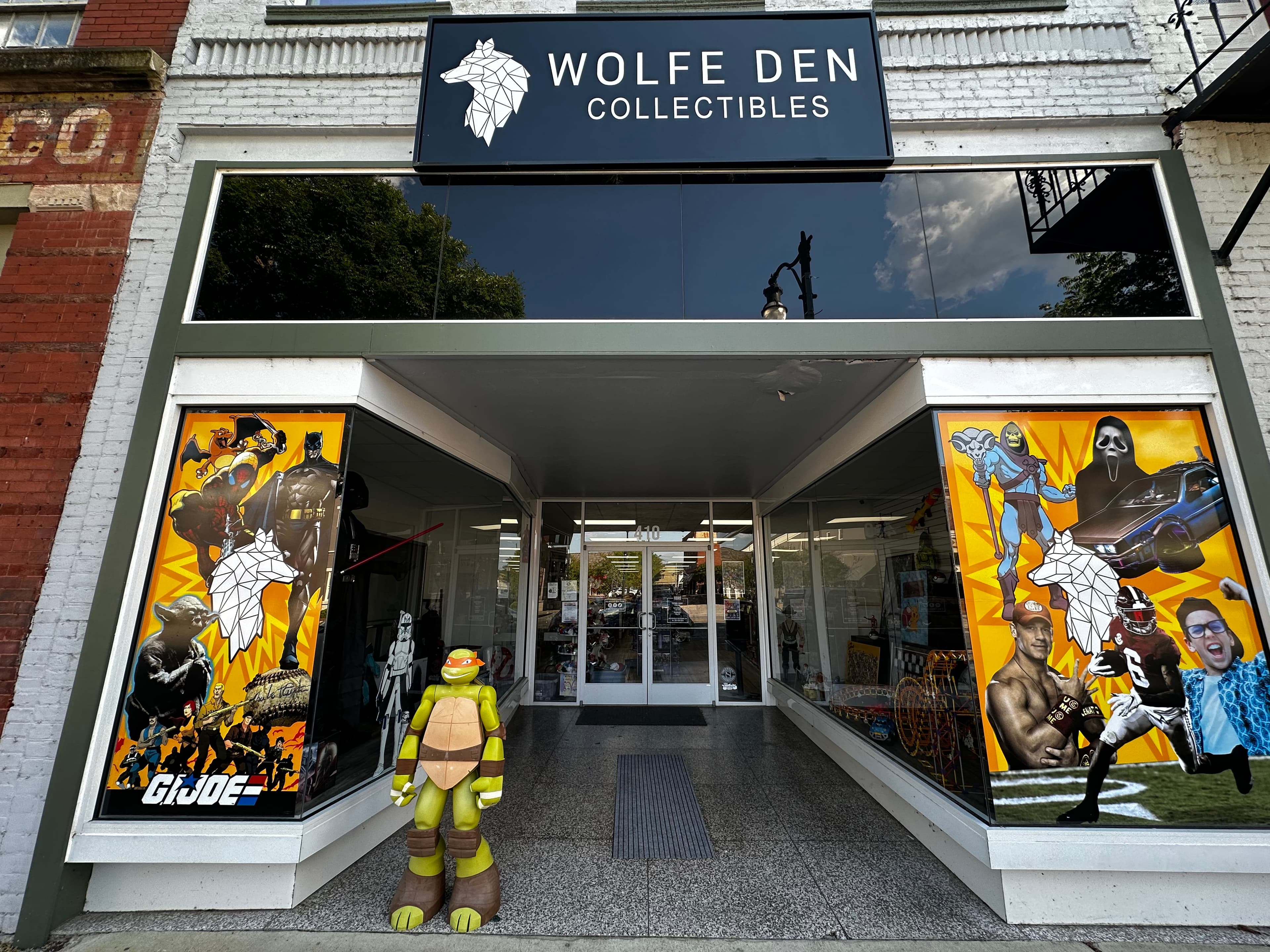 Wolfe Den Collectibles storefront featuring pop culture window graphics and a life-sized Ninja Turtle statue.